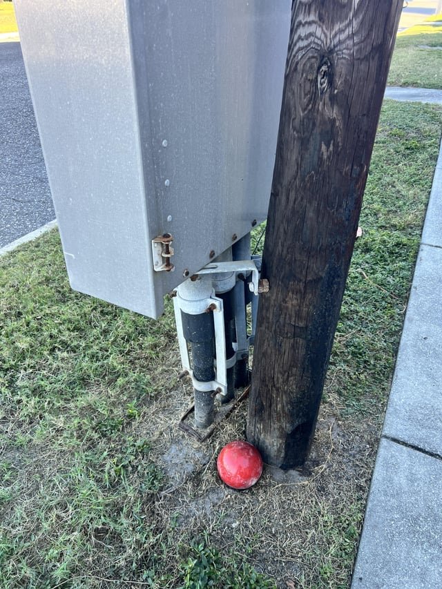​This round, red rubbery object that is attached to the ground. It is next to a utility box of some unidentified type.