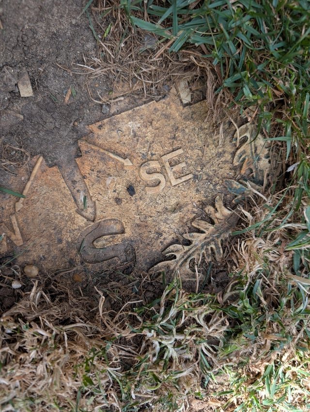 ​What is this thing buried in my back garden? The exposed part is about the size of my hand. I think it’s made of plastic. Assume it’s some sort of marker but would like to know if I can remove it as the grass around it dies. East Anglia, UK.